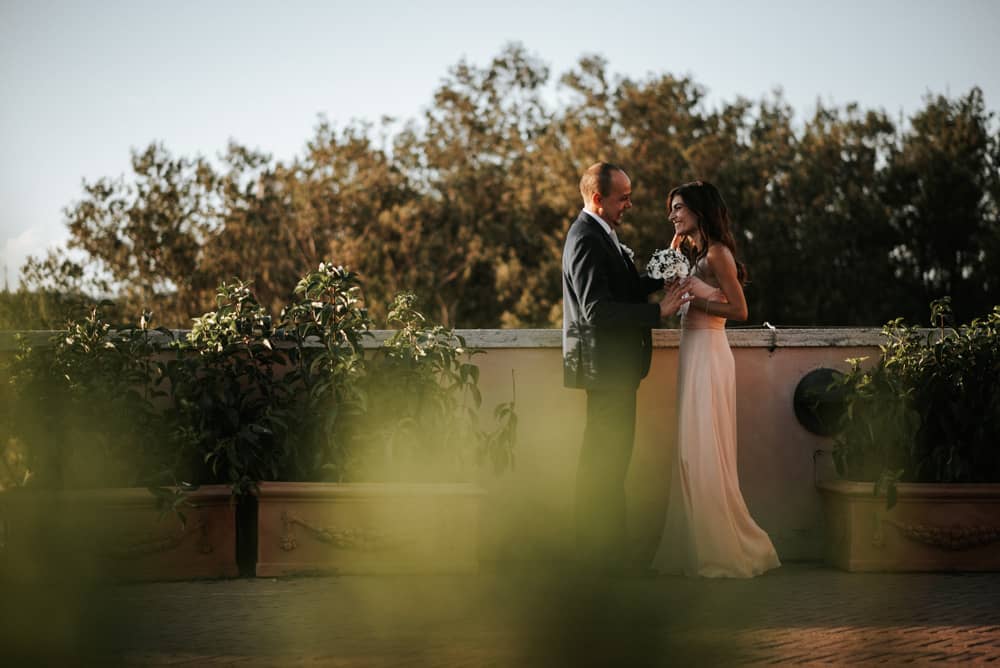 Sposi che si guardano con amore, con la sposa che tiene in mano un bouquet, sullo sfondo delle Terrazze Caffarelli a Roma.