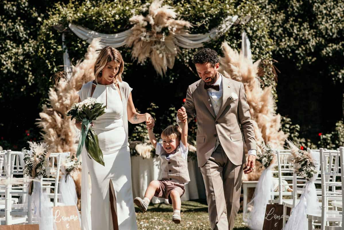Momento di gioia con il bambino e l’allestimento boho chic al matrimonio ai Castelli Romani Sposi con il loro bambino sorridenti mentre escono dal rito civile in una location ai Castelli Romani, circondati da un allestimento boho chic, prima del lancio del riso.