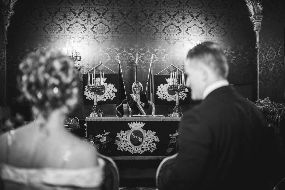 fotografo-matrimonio-roma-rito-civile-spontaneo-zero-stress Sposi di spalle durante la cerimonia di rito civile in un Comune di Roma con celebrante, scatto in stile reportage spontaneo Metodo Zero Stress.