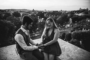 Coppia, Francesco e Loise, seduti sulla balconata della terrazza del Giardino degli Aranci a Roma durante una proposta di matrimonio, con l'anello in primo piano e vista panoramica di Roma sullo sfondo.